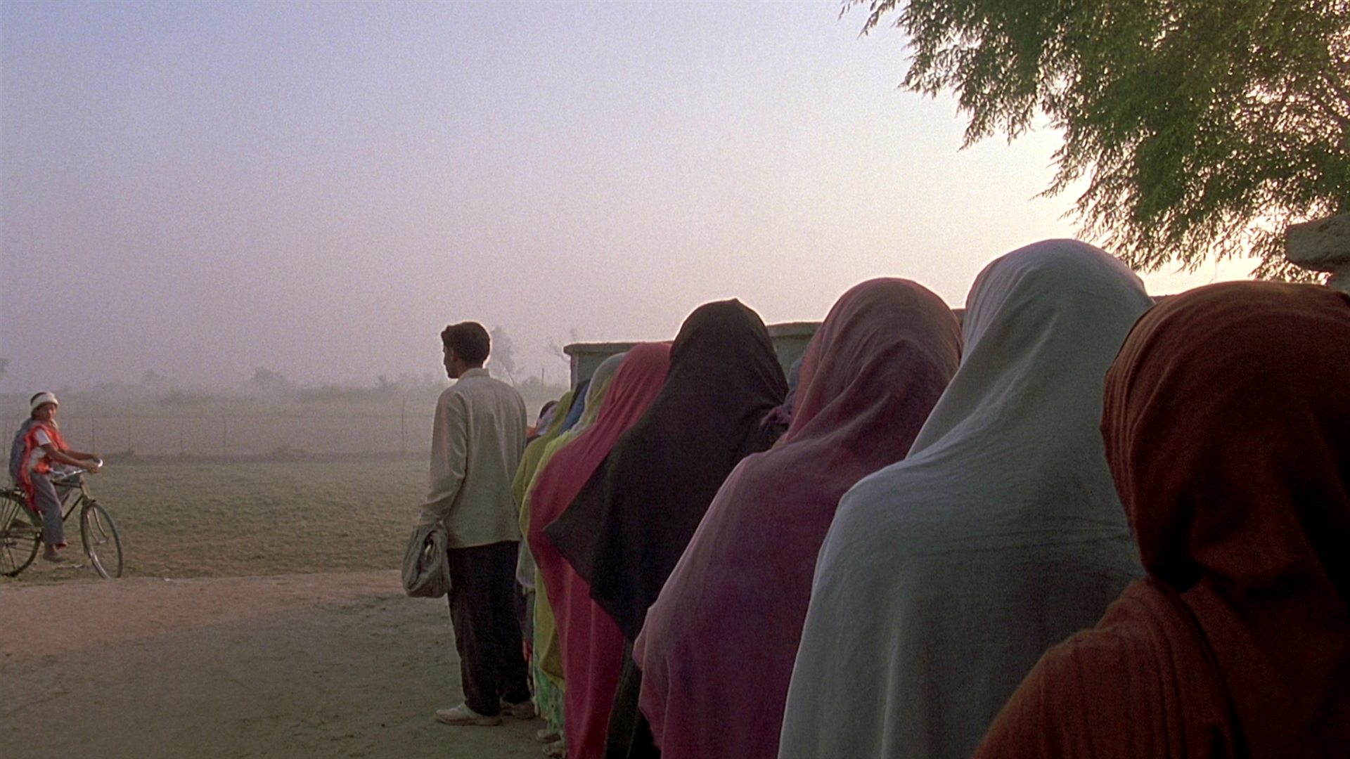 Early morning: a line of people, seen from behind, waiting outside a village school in mist.