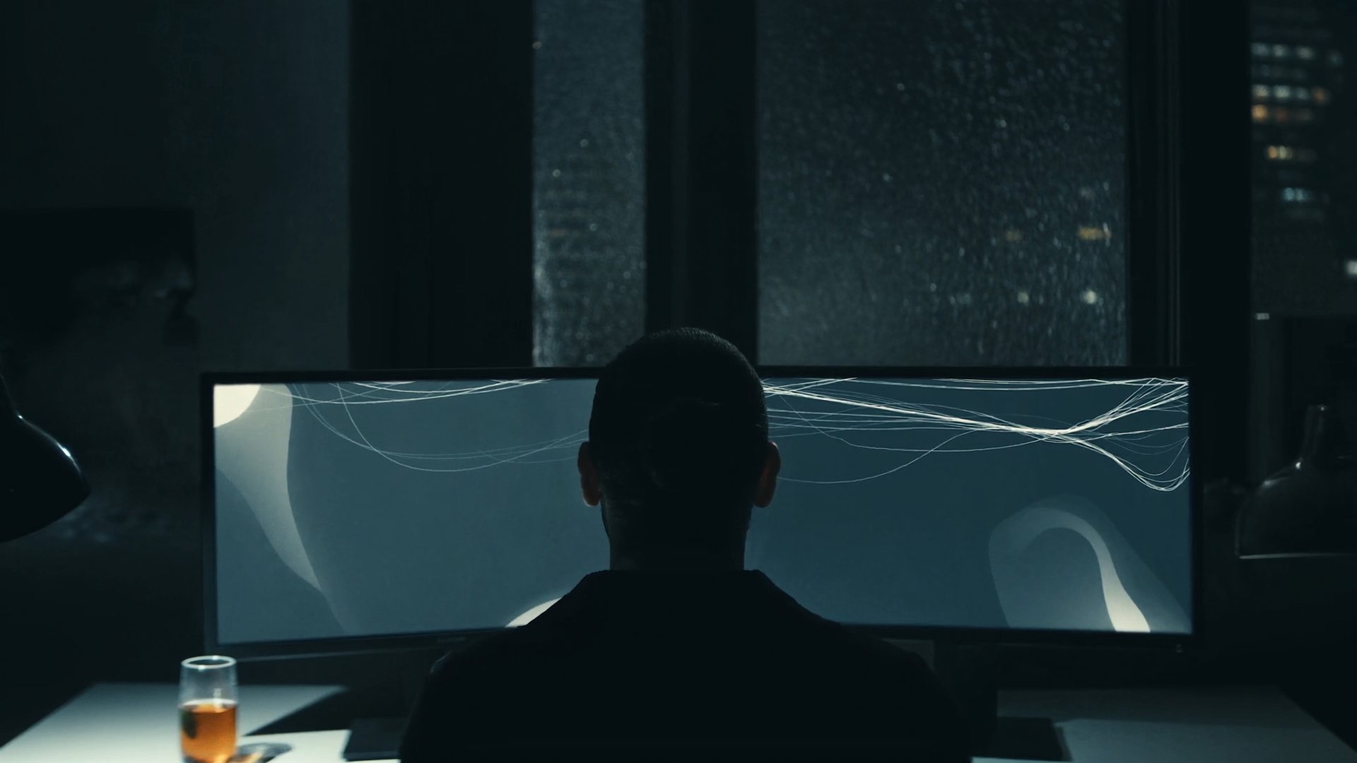 An analyst seen from behind at night, working at two monitors showing abstract lines of light.